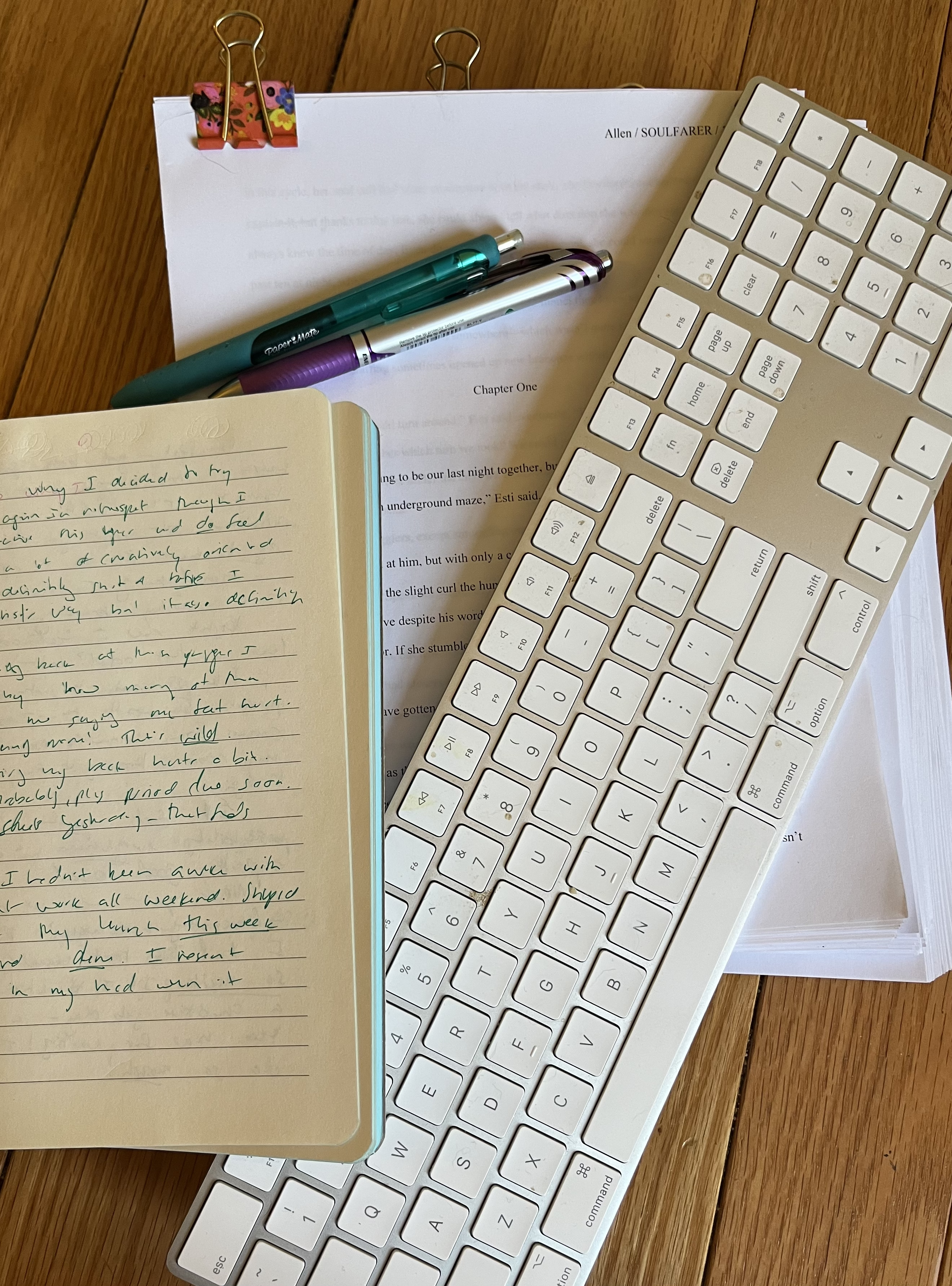 A collage on a wooden floor: A thick packet of papers (a printed manuscript), pens, a keyboard, and an open notebook